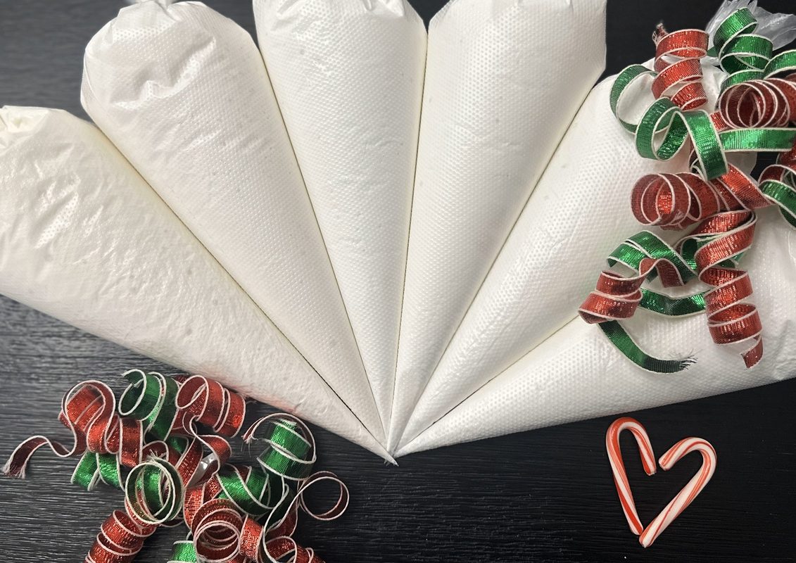Six tipless icing bags filled with white royal icing fanned out with red and green curling ribbon and a heart made from two mini candy canes