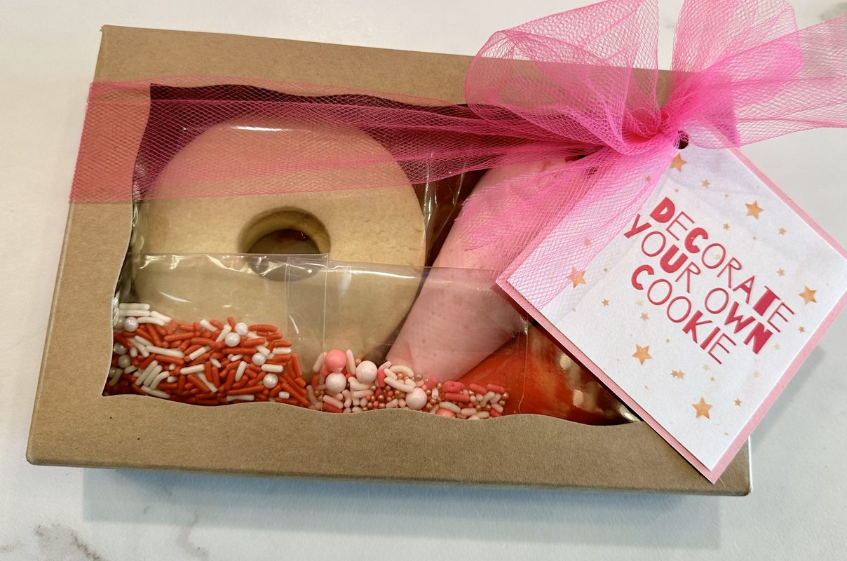 A single DIY cookie kit in a window box with a donut cookie, icing bag and sprinkles, tied with a tulle bow that wraps all the way around the box and a Decorate Your Own Cookie tag in the corner