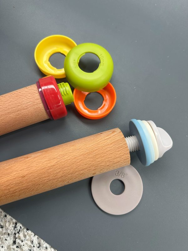 Kathy's two adjustable rolling pins side by side — the colored ring style with its rings popped off at the top, and the screw-in style she recommends at the bottom with its thickness discs on the threaded end