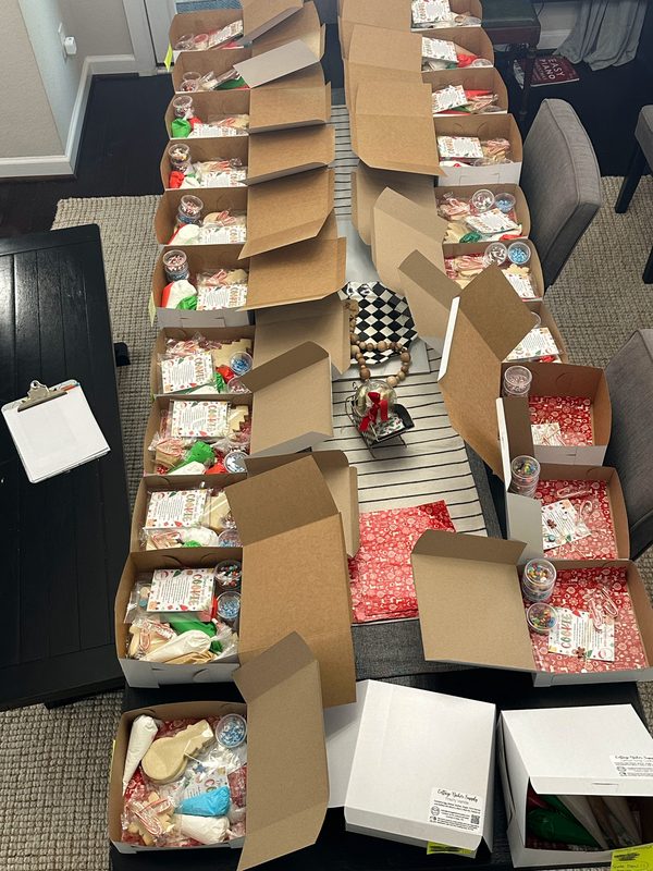 Rows of kit boxes: Rows of DIY cookie kit boxes lined up on a dining table being packed with cookies, icing bags, sprinkle cups and instruction sheets for customer orders