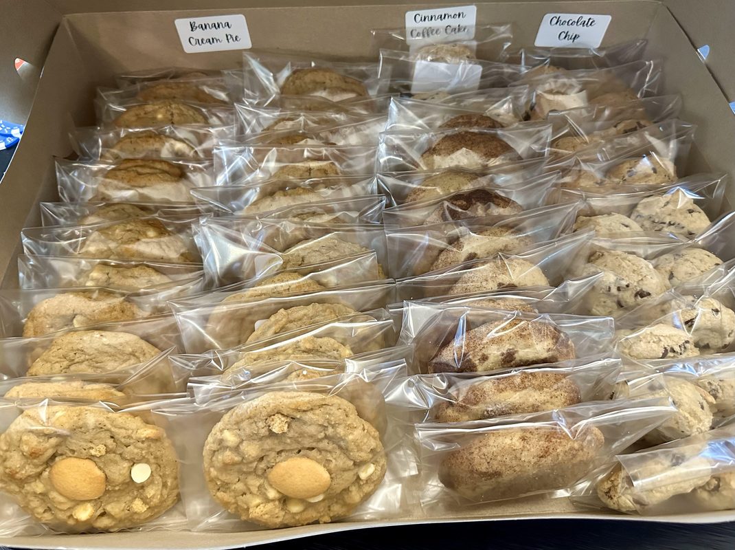 A bakery box full of individually bagged and sealed drop cookies with flavor labels — banana cream pie, cinnamon coffee cake, and chocolate chip