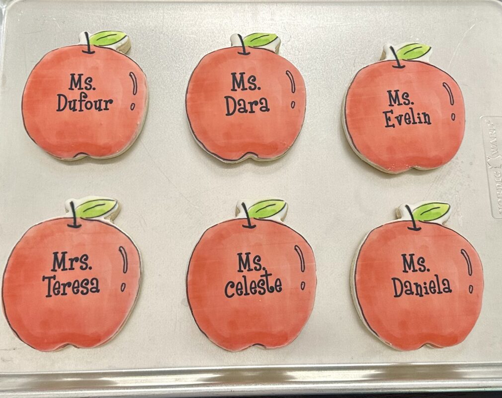 Six bright red printed apple sugar cookies on a sheet pan, personalized with teacher names like Ms. Dufour and Ms. Dara