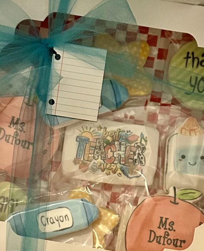 A professional white bakery window box holding individual poly-bagged teacher cookies lined with red checkered paper and a turquoise ribbon bow