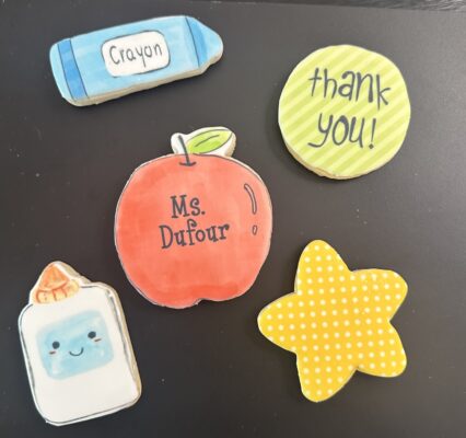 A flat-lay arrangement of five teacher appreciation sugar cookies including a personalized apple, a blue crayon, glue bottle, star, and thank you token