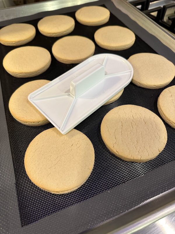 Ateco fondant smoother, Kathy's cookie iron, resting on fresh round sugar cookies baked on a Cookie Countess perforated mat on a Nordic Ware half sheet pan