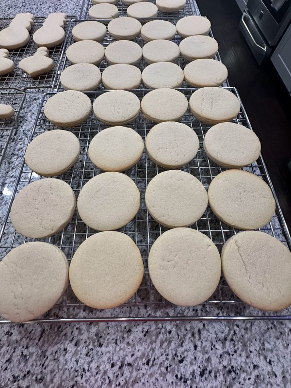 Rows of large round sugar cookies cooling on racks — a full batch of elf pizza cookie bases ready for printing