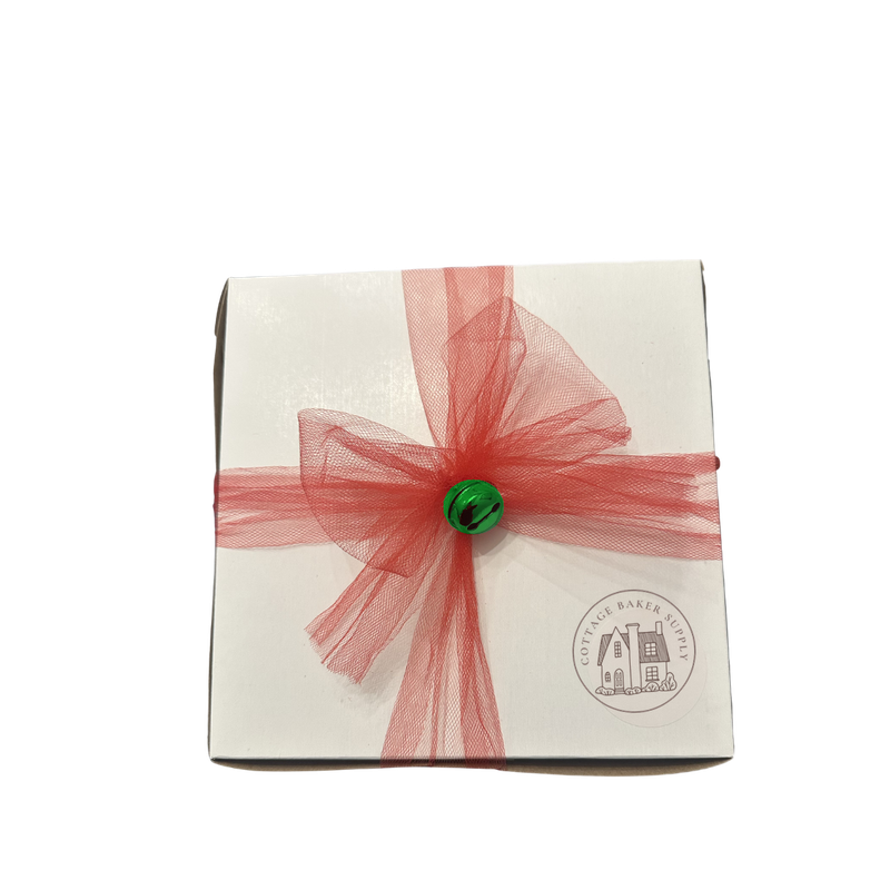 Kathy's white DIY cookie kit box tied with red tulle that wraps around all four sides, a bow and jingle bell at the center, and a round sticker in the bottom right corner